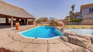 Ein Pool mit Steinrand, ein strohgedecktes Gebäude und ein Steinhaus mit Ziegeldach.