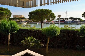 A waterfront view with boats, a tree, and a building in the background. - Cozy Coastal Flat Facing the Port (Lignano Sabbiadoro)