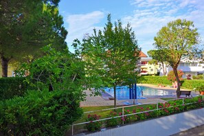 A swimming pool surrounded by lush greenery and a building in the background. - Cozy Coastal Flat Facing the Port (Lignano Sabbiadoro)