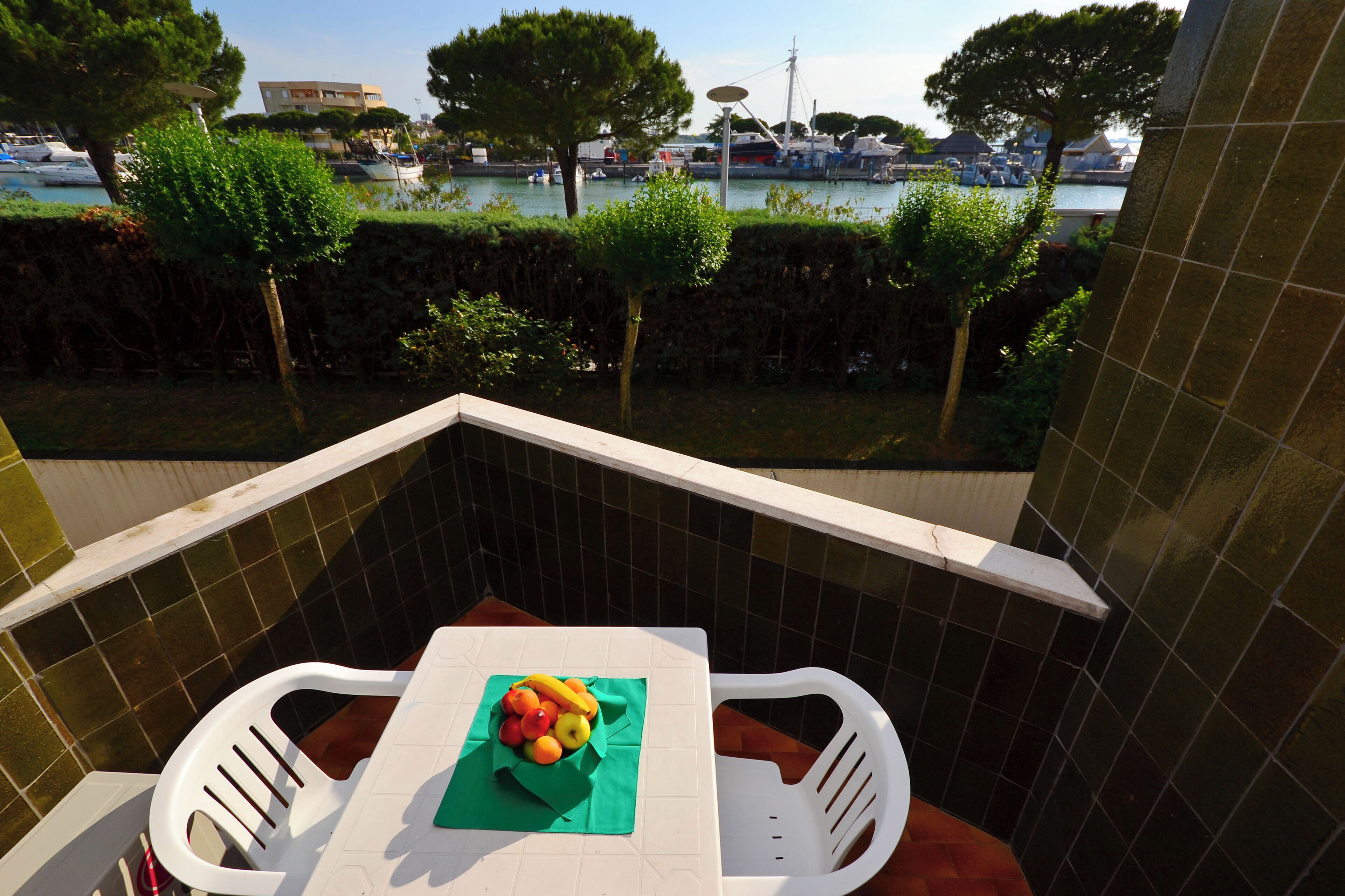 Un balcon avec un mur recouvert de carreaux, une table sur laquelle se trouve un panier de fruits, et une vue sur une marina avec des bateaux et des arbres.