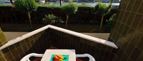 Un balcon avec un mur recouvert de carreaux, une table sur laquelle se trouve un panier de fruits, et une vue sur une marina avec des bateaux et des arbres.