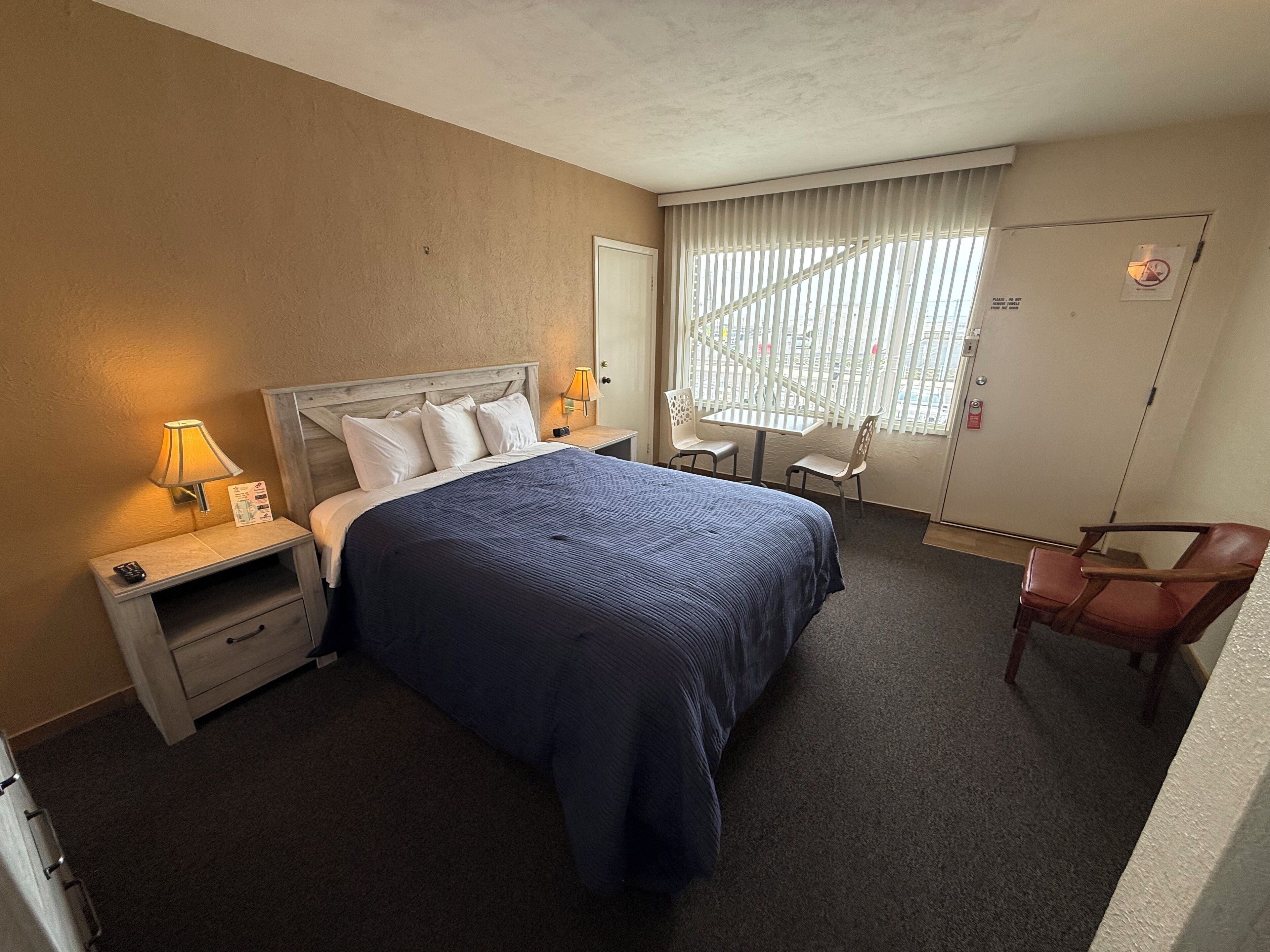 Habitación de hotel con cama, mesitas de noche, una silla, una mesita pequeña y una ventana con persianas.