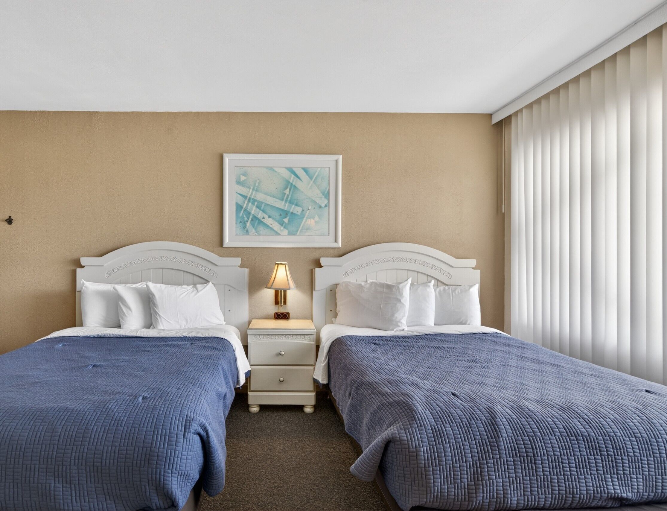 Habitación de hotel con dos camas, una mesita de noche con lámpara, un cuadro enmarcado en la pared y una ventana con persianas.