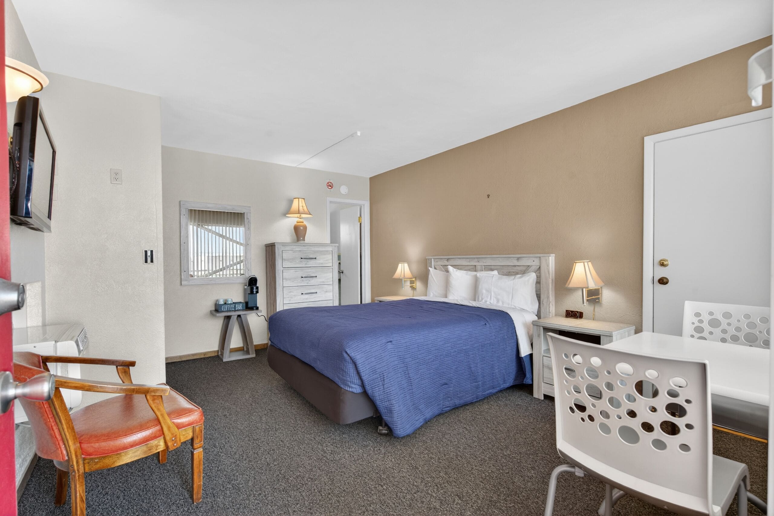 Ein Hotelzimmer mit Bett, Schreibtisch, Stuhl, Lampe, Kommode und einer Tür.