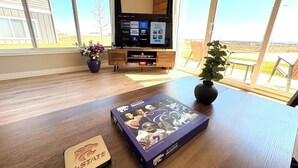 Ein Wohnzimmer mit Holzboden, einem Fernseher auf einem Schrank, einem Tisch mit einem Buch und einem Untersetzer sowie einem Blick ins Freie durch große Fenster.