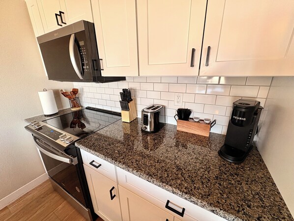 A modern kitchen with white cabinets, a granite countertop, a built-in oven, a microwave, a toaster, and a Keurig coffee maker. - Colbert Hills Retreat + K-State Stay & Hilltop Views! (Manhattan)