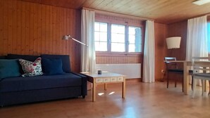 A wooden-paneled room with a sofa, a small table, a chair, and a window with curtains.
