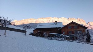 A wooden chalet with a sloped roof, surrounded by snow-covered hills and mountains.