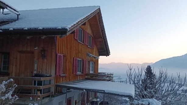 A wooden house with a balcony, surrounded by snow-covered landscape and mountains in the background.