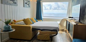 Ein Zimmer mit Bett, Schreibtisch, Stuhl, einem kleinen Tisch mit einer Vase und Blick aufs Meer.