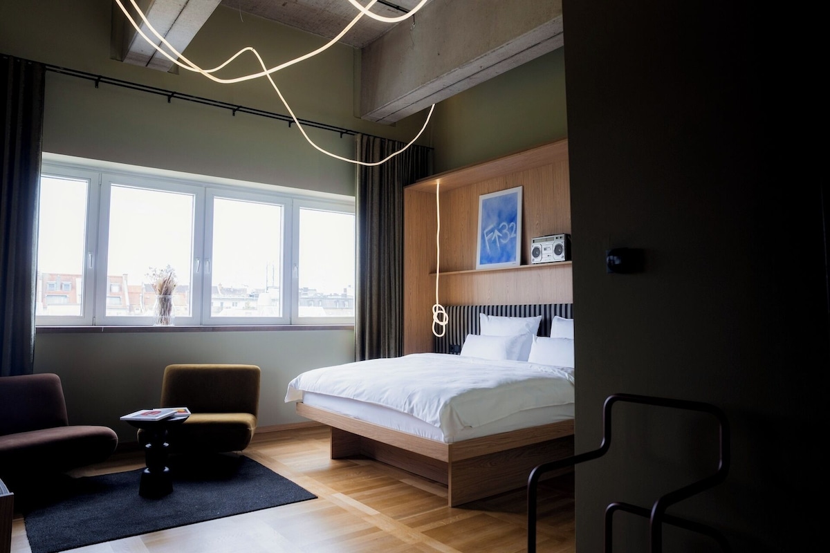A modern hotel room with a large bed, a desk, a chair, and a view of the city through large windows.