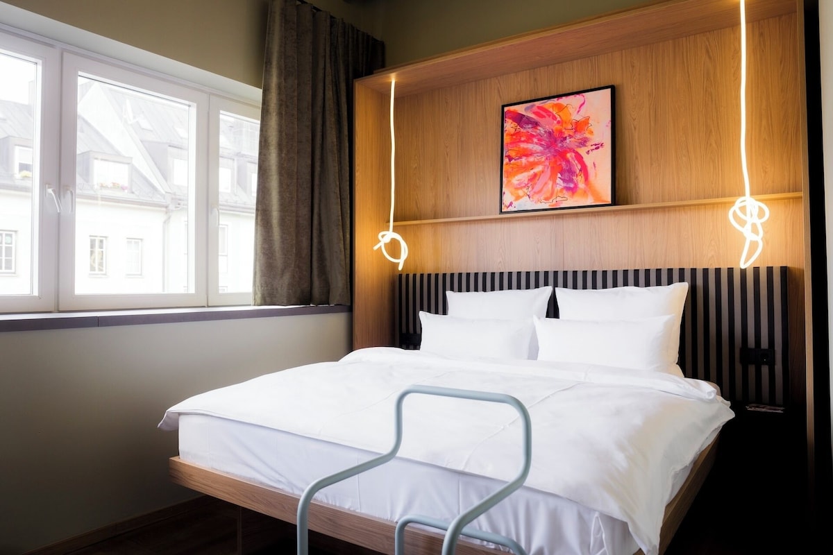 A hotel room with a bed, a window, a painting, and a bedside lamp.