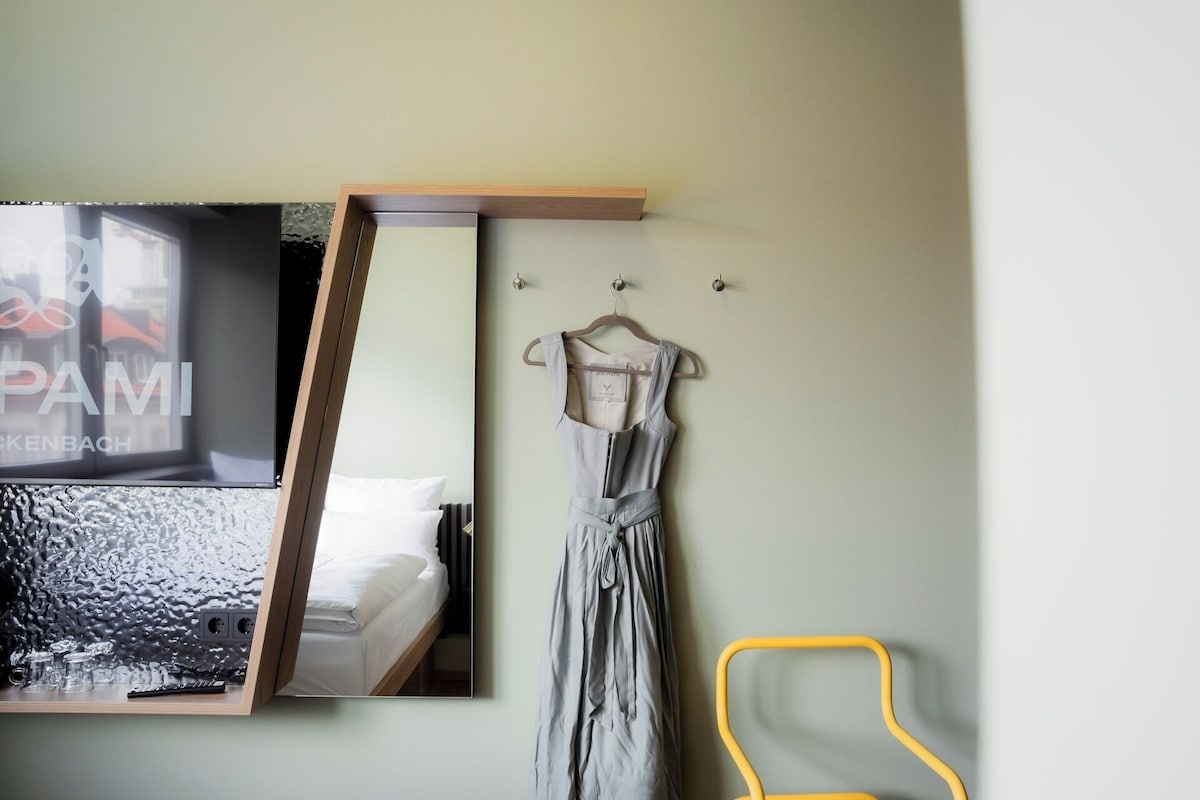 A room with a bed, a mirror, a hanging dress, and a yellow chair.
