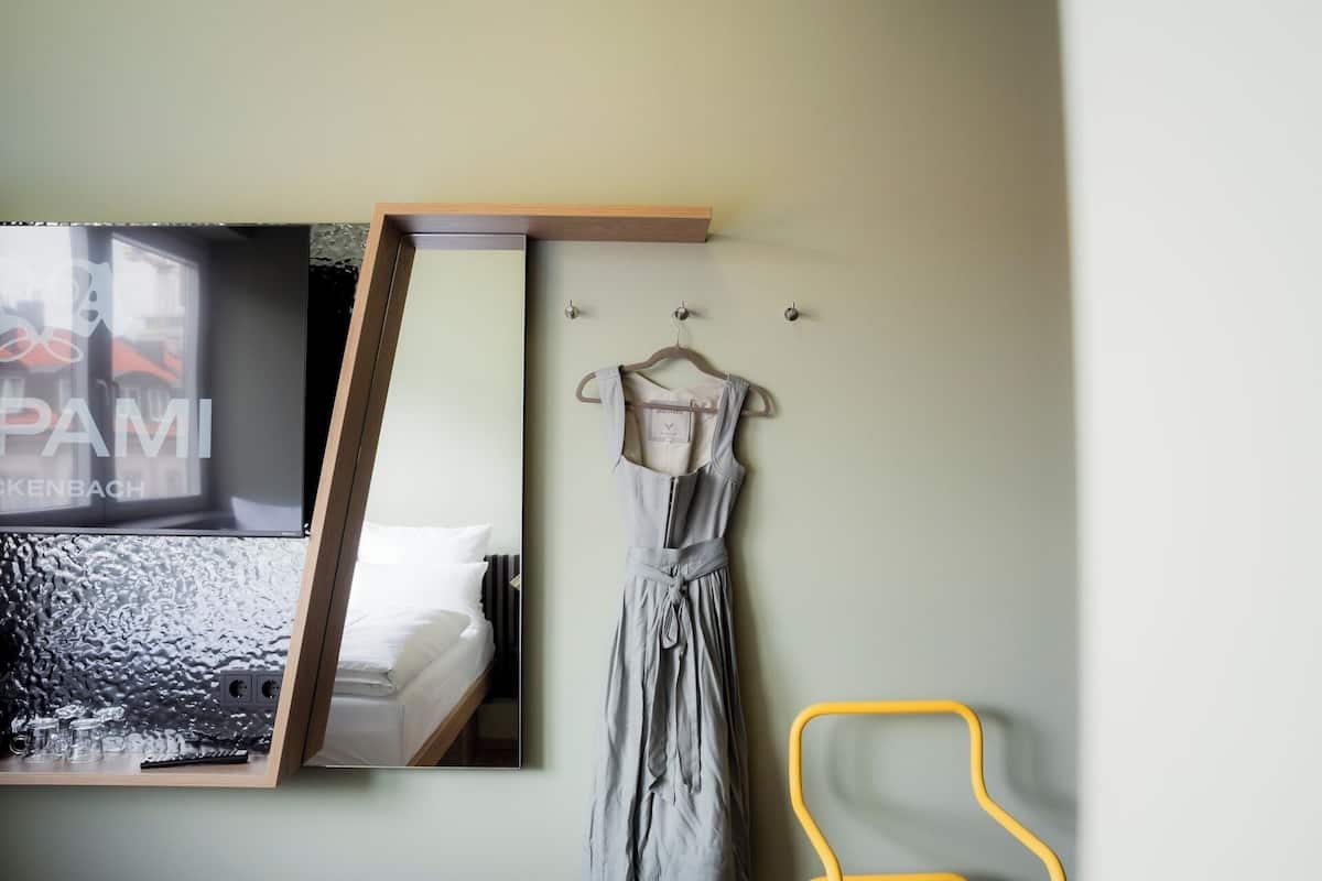 Una habitación con cama, espejo, un vestido colgado y una silla amarilla.