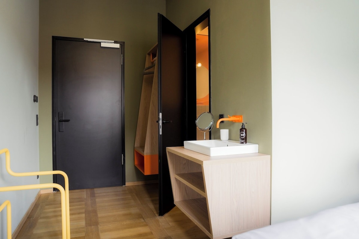 A hotel room with a wooden floor, a bed, a bathroom with a sink and mirror, and a door leading to another room.