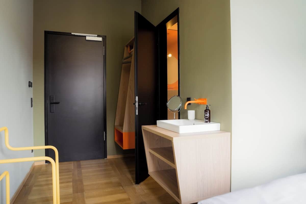 Una habitación de hotel con piso de madera, una cama, un baño con lavamanos y espejo, y una puerta que da a otra habitación.