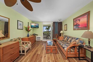 A living room with a sofa, coffee table, and wicker furniture. There is a television mounted on the wall and a window with curtains. - Summer Deal Kona Ocean View Studio Walk to Beaches Dining and Shops (Kailua-Kona)