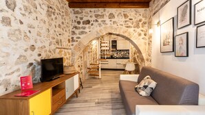 A stone-walled room with a wooden ceiling, a television on a yellow cabinet, a gray sofa, and a kitchen visible through an archway. - La Naca by Wonderful Italy (Syracuse)