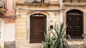A building with a brown door, a potted plant, and a stone-paved ground. - La Naca by Wonderful Italy (Syracuse)