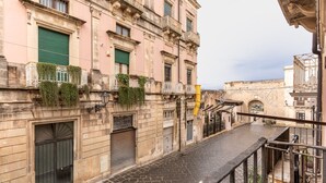 A narrow street with a building featuring a green shutter, a balcony with plants, and a metal railing. - La Naca by Wonderful Italy (Syracuse)