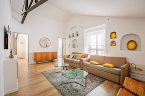 Ein geräumiges Wohnzimmer mit Holzboden, einem Glastisch, einer Couch mit Kissen, einer Holzkommode und einem großen Fenster.