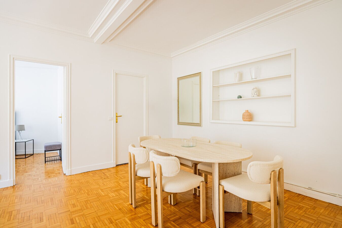 Een eetruimte met een ronde tafel en zes stoelen, een deur naar een andere kamer, en een wandplank met decoratieve objecten.