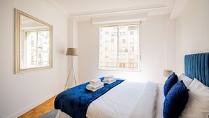 Ein Hotelzimmer mit einem Bett, einem großen Spiegel, einer Stehlampe und einem Fenster mit Blick auf Gebäude.