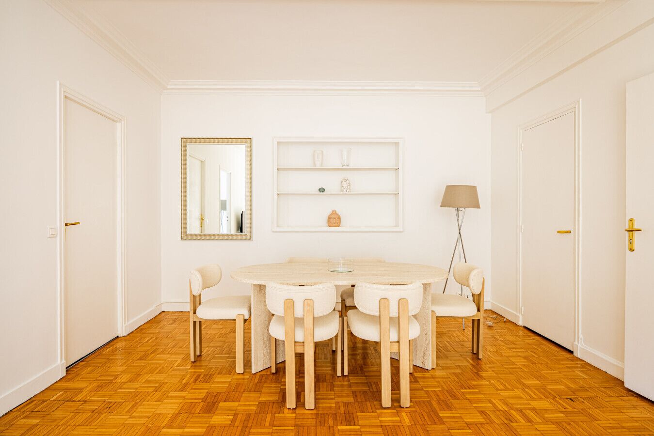 Una habitación con una mesa redonda, seis sillas, un espejo, estanterías, una lámpara y puertas blancas.