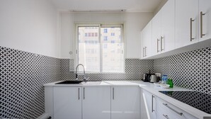Eine Küche mit weißen Schränken, einer schwarz-weißen Spritzschutzwand, einem Fenster mit Jalousien und einer Arbeitsplatte mit Wasserkocher und Flaschen.