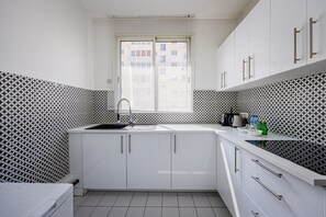A kitchen with white cabinets, a black and white patterned backsplash, a window with blinds, and a countertop with a kettle and bottles. - Premium 1-Bedroom - Trocadéro - Eiffel Tower - Paris 16 (Paris)