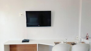 Una camera d'albergo con una TV a schermo piatto montata a parete, una scrivania in legno con un portatile e un'area ristoro con un tavolo bianco apparecchiato per due.