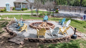 A circular fire pit area with Adirondack chairs, a brick fire pit, and a house in the background.