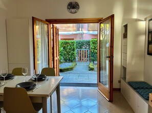 Un coin repas avec une table dressée pour deux personnes, une horloge au mur et une vue sur un jardin par une porte ouverte.
