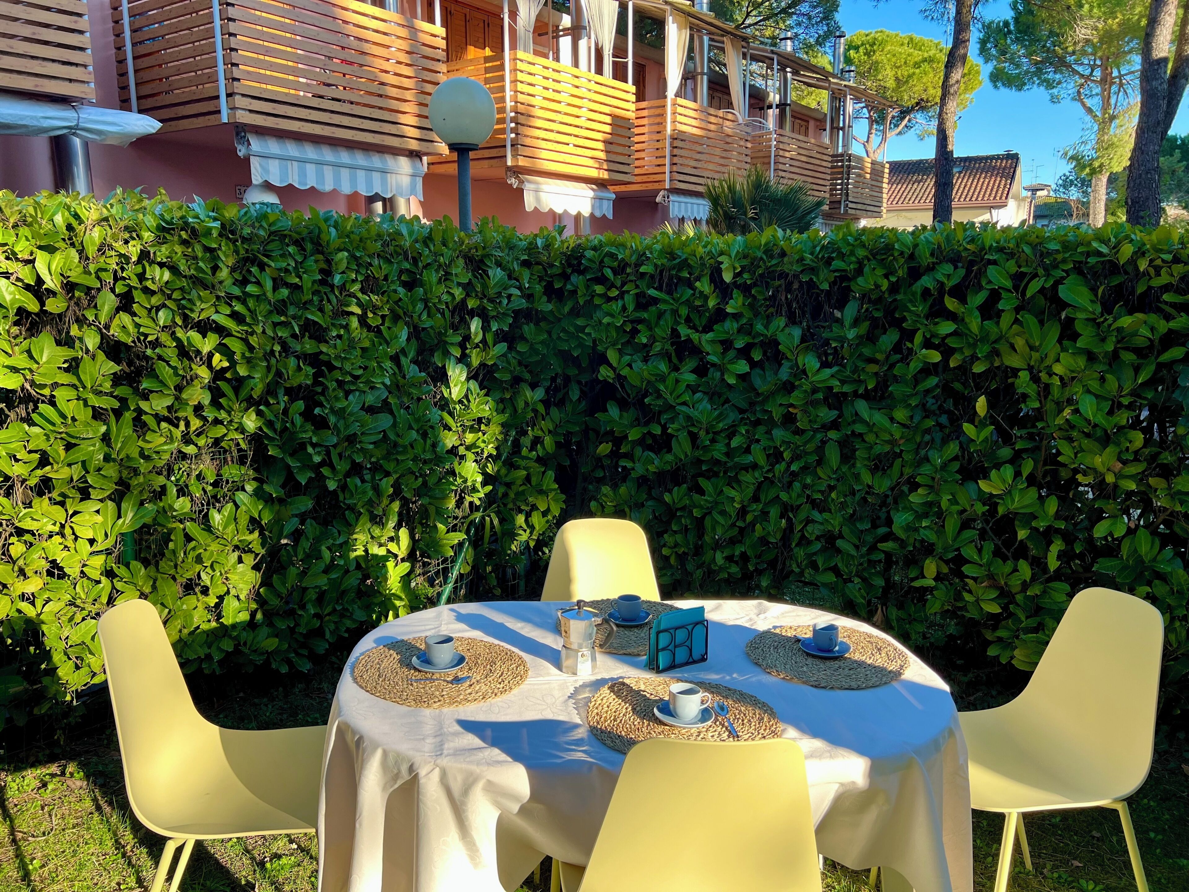 An outdoor dining area with a table set for four, surrounded by chairs, and a hedge in the background.