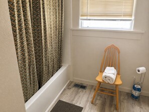 A bathroom with a shower curtain, a wooden chair with a towel, a toilet paper holder, and a vent.