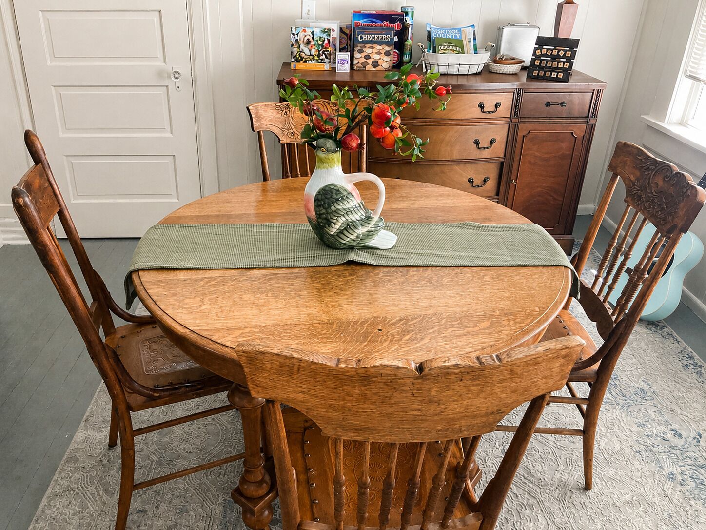 A wooden dining table with chairs, a vase with flowers, and a checkers board on a wooden cabinet.