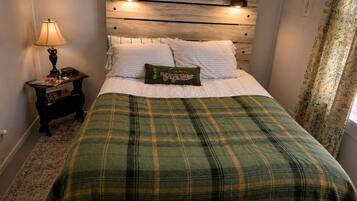 A bedroom with a wooden headboard, a green plaid bedspread, a nightstand with a lamp, and a floral curtain.