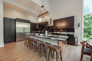 Une cuisine moderne avec des meubles de couleur sombre, un réfrigérateur en acier inoxydable, un grand îlot central doté de tabourets et un luminaire suspendu.
