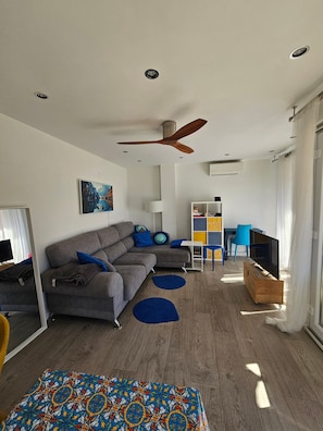 Un salon moderne avec un canapé gris, des coussins bleus, une table basse en bois, un téléviseur posé sur un meuble en bois et un ventilateur de plafond.