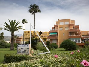Cartell per 'JARDINES DEL MAR' en una zona verda amb palmeres i un edifici amb balcons.