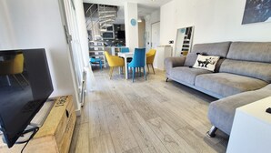 A modern living room with a grey sofa, a TV, and a dining area with blue chairs. - Apartment 'Duplex Jardines Del Mar' with Sea View, Wi-Fi and Air Conditioning (Salobreña)