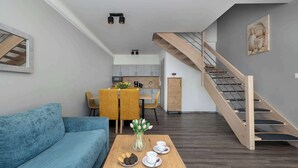 Ein modernes Wohnzimmer mit einer Couch, einem Couchtisch mit Keksen und Tassen, einem Essbereich mit Stühlen und einer Blumenvase, einer Küche im Hintergrund und einer Holztreppe.