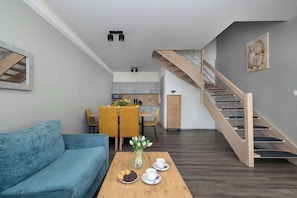 Ein modernes Wohnzimmer mit einer Couch, einem Couchtisch mit Keksen und Tassen, einem Essbereich mit Stühlen und einer Blumenvase, einer Küche im Hintergrund und einer Holztreppe.