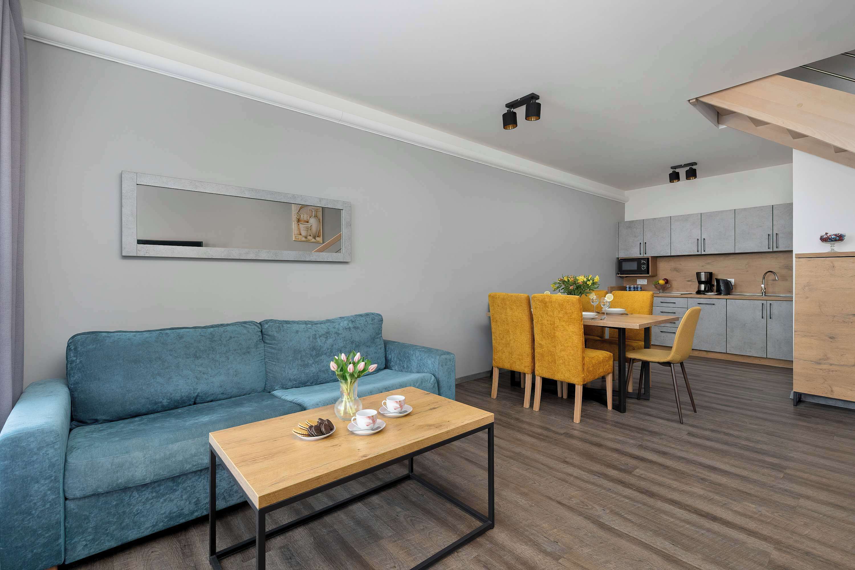 A bright open-plan living room features a comfortable teal blue sofa, a wooden coffee table, and a dining area with vibrant yellow chairs.