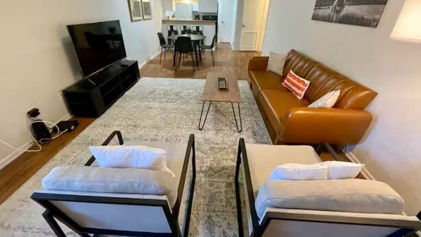 Una sala de estar con televisor, sofá y dos sillones, un comedor con sillas y mesa, y una cocina al fondo.