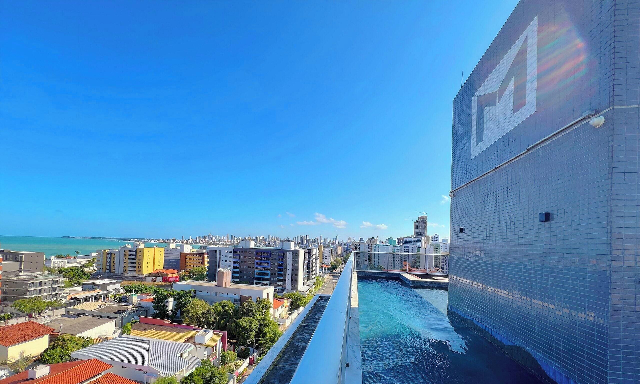 A high-rise building with a large window, a swimming pool, and a cityscape with buildings and a distant coastline.