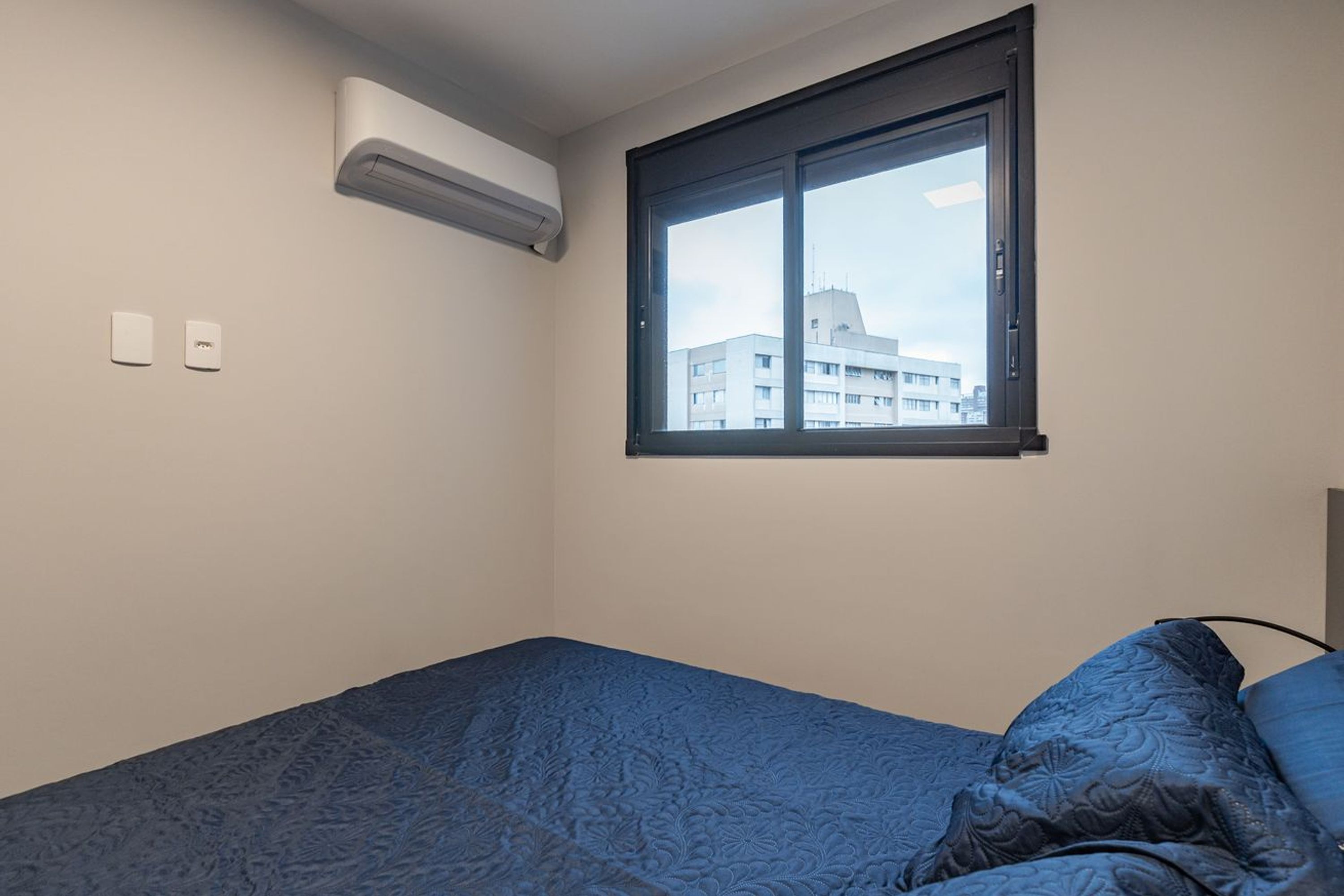 Une chambre avec un lit simple, une fenêtre donnant sur des immeubles, un appareil de climatisation et deux interrupteurs fixés au mur.