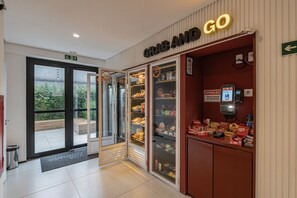 Ein Hotel Eingang mit einem 'GRABAND GO' Schild, einer Glastür, einem Empfangsbereich mit Snacks und einem Schild für 24-Stunden-Service.