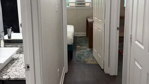 Ein schmaler Flur mit einem Badezimmer links, einem Schlafzimmer mit Fenster und Klimaanlage in der Mitte und einer Tür zu einem weiteren Raum rechts.