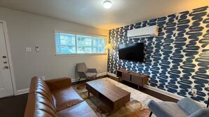 Ein Wohnzimmer mit braunem Ledersofa, einem Holztisch, einem grauen Sessel, einer Wand mit Muster und einem an der Wand befestigten Flachbildfernseher.